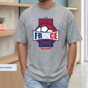 May include: An athletic heather gray short-sleeve t-shirt featuring a graphic design with a soccer ball, the text "WORLD CUP FRANCE 2026," and two small stars. The design uses blue, red, and white colors centered on the chest of the garment.