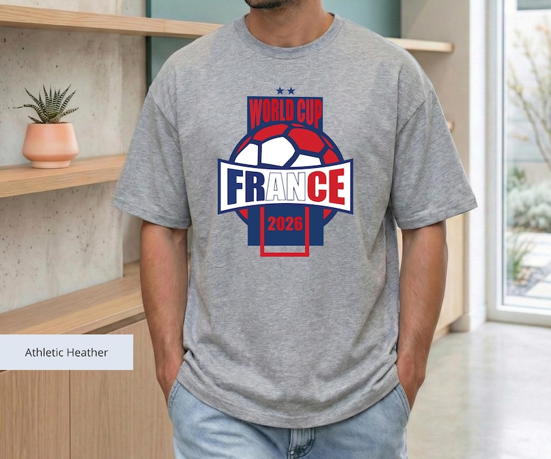 May include: An athletic heather gray short-sleeve t-shirt featuring a graphic design with a soccer ball, the text "WORLD CUP FRANCE 2026," and two small stars. The design uses blue, red, and white colors centered on the chest of the garment.