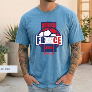 May include: A steel blue short-sleeved t-shirt featuring a graphic print that includes a soccer ball, the text "WORLD CUP", "FRANCE", and "2026" in red, white, and blue colors, with two small stars positioned above the text.