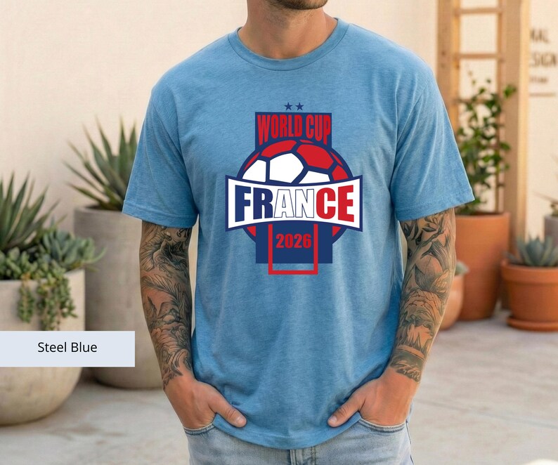 May include: A steel blue short-sleeved t-shirt featuring a graphic print that includes a soccer ball, the text "WORLD CUP", "FRANCE", and "2026" in red, white, and blue colors, with two small stars positioned above the text.