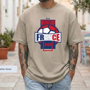 May include: A heather stone colored t-shirt featuring a graphic design with a soccer ball, the text "WORLD CUP FRANCE 2026", and two small stars. The design uses red, white, and blue colors, centered on the chest of the short-sleeved garment.