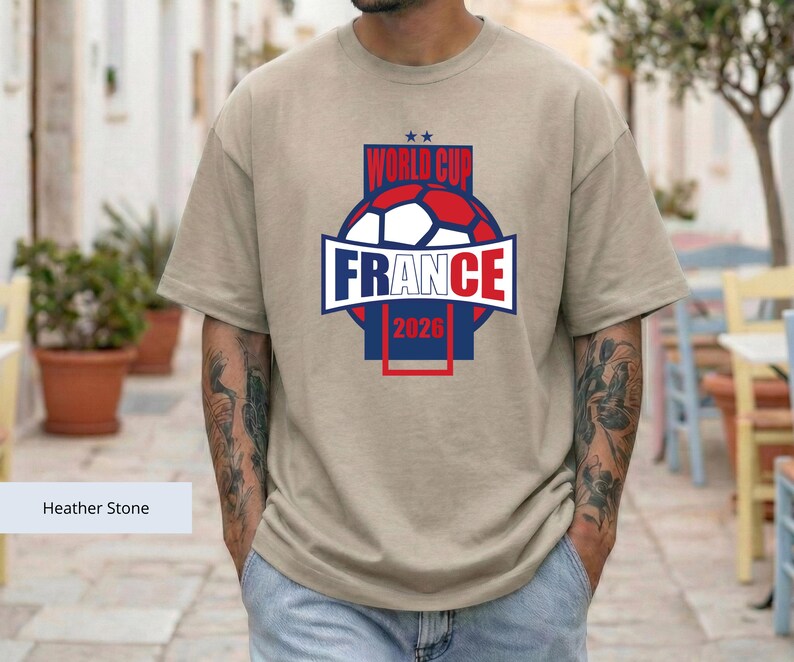 May include: A heather stone colored t-shirt featuring a graphic design with a soccer ball, the text "WORLD CUP FRANCE 2026", and two small stars. The design uses red, white, and blue colors, centered on the chest of the short-sleeved garment.