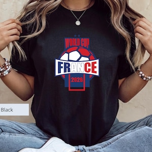 May include: A black short-sleeve t-shirt featuring a graphic design with a soccer ball and the text "WORLD CUP FRANCE 2026" in red, white, and blue colors. The design includes a central banner with the word "FRANCE" and the year "2026" below.