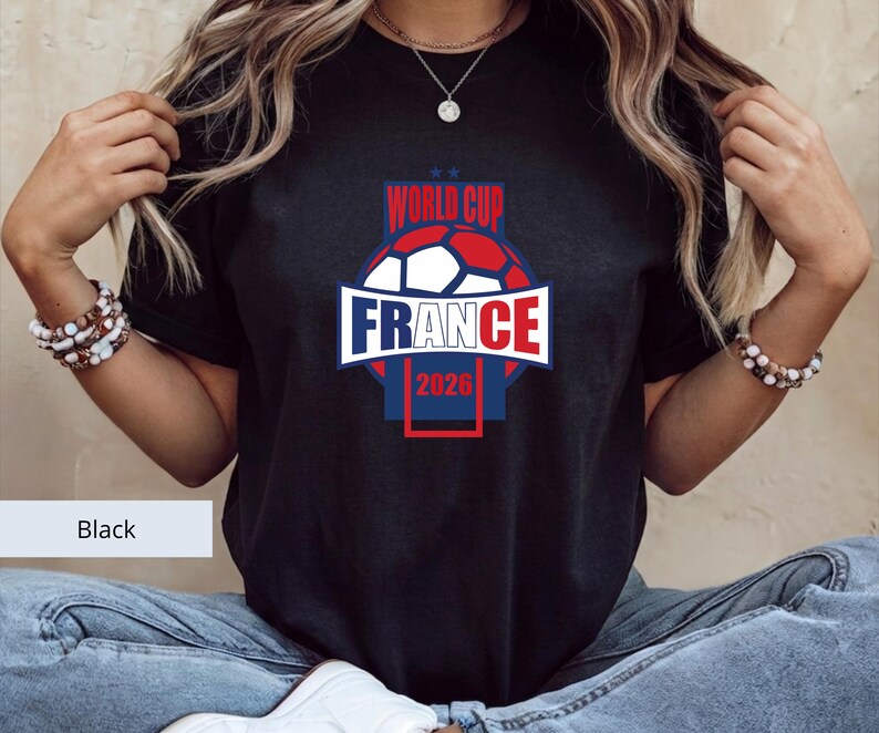 May include: A black short-sleeve t-shirt featuring a graphic design with a soccer ball and the text "WORLD CUP FRANCE 2026" in red, white, and blue colors. The design includes a central banner with the word "FRANCE" and the year "2026" below.