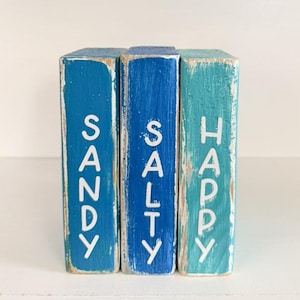 May include: Three wooden blocks painted in shades of blue, with the words "SANDY", "SALTY", and "HAPPY" in white lettering. The blocks have a distressed, weathered appearance, suggesting a coastal or beach theme.