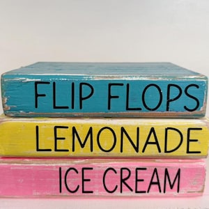 May include: Three stacked, rectangular wooden blocks painted in turquoise, yellow, and pink. Each block has black lettering: "FLIP FLOPS," "LEMONADE," and "ICE CREAM." The blocks have a distressed, rustic appearance.