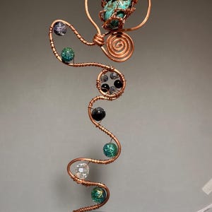 May include: A decorative copper wire sculpture featuring a central turquoise stone wrapped in wire, accented with a spiral design and a series of multi-colored beads including green, black, clear, and speckled spheres arranged along a wavy wire stem.