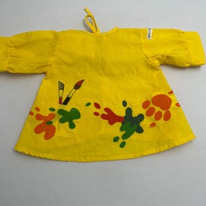 May include: A bright yellow long-sleeved smock featuring two front pockets, a tie closure at the neckline, and colorful paint splatter graphics in red, orange, and green. One pocket displays two paintbrushes, while the other shows a paw print.