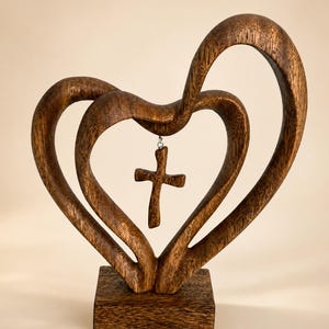 Könnte beinhalten: Eine dunkelbraune Holzskulptur mit zwei ineinander verschlungenen Herzformen auf einem rechteckigen Sockel. Ein kleines Holzkreuz hängt an einem silberfarbenen Metallring, der oben in der Mitte des inneren Herzens befestigt ist.