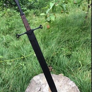 May include: A long sword with a matte black blade and a matching black crossguard. The handle features a brown grip with visible rivets and a triangular pommel at the end. The sword is positioned vertically with the tip resting on a wooden stump.
