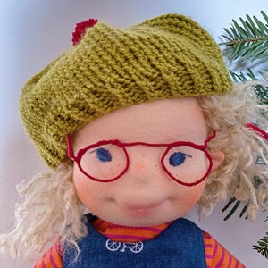 Puede incluir: Una muñeca hecha a mano con cabello rubio rizado que lleva una boina de punto verde con un pompón rojo, gafas rojas y un vestido de mezclilla azul con rayas naranja y blancas.