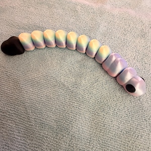 Puede incluir: Un juguete de oruga articulado y colorido. La cabeza es negra y el cuerpo transita por tonos pastel de amarillo, azul y rosa. El juguete está diseñado con secciones segmentadas, lo que permite un movimiento y una postura flexibles.