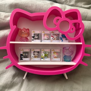 May include: A vibrant pink Hello Kitty-shaped display shelf holding several Funko Pop figures in their boxes. The shelf has two white shelves and is supported by two white legs. The figures feature various Hello Kitty characters.