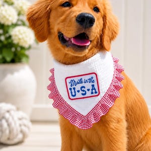 Op de afbeelding: Een golden retriever puppy draagt een witte driehoekige bandana voor huisdieren met een rood-witte gingham ruche rand. Het midden van de bandana heeft een rechthoekige patch met een rode rand en de blauwe tekst Made in the U.S.A.