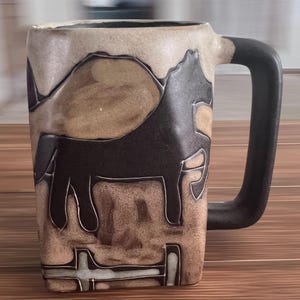 Peut inclure: Un mug en céramique de forme carrée présentant la silhouette d'un cheval brun foncé sur un fond beige texturé. Le mug possède une anse épaisse brun foncé et une base plate. La surface présente une finition mate avec des contours sculptés.