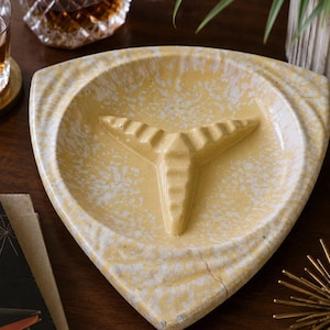 Puede incluir: Un cenicero triangular de cerámica con un esmalte moteado en amarillo y blanco. El centro del plato contiene una estructura de soporte elevada de tres puntas con detalles acanalados, diseñada para sujetar objetos en la base del recipiente.