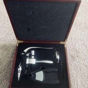 Puede incluir: Un juego de accesorios para vino presentado en una caja de madera de color marrón oscuro con tapa abatible. El set incluye un abridor de metal plateado, un tapón, un cortacápsulas y un vertedor, organizados en espuma negra a medida.