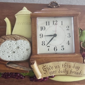 May include: A wall clock with a wooden frame featuring a cream-colored teapot, a loaf of bread, a knife, and clusters of grapes. A banner at the bottom displays the text, Give us this day our daily bread. The clock face has black numbers.