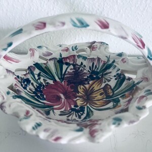 May include: A white ceramic basket with a scalloped, openwork rim and a curved handle. The interior features a hand-painted floral design in shades of red, yellow, and green. Three-dimensional ceramic flowers are attached to the handle base.
