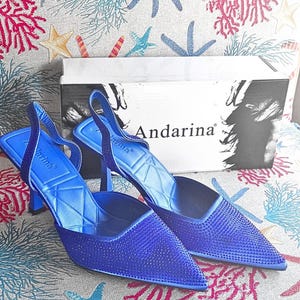 Puede incluir: Un par de zapatos de tacón alto azules con punta y correas traseras. Los zapatos están cubiertos de pequeños adornos brillantes. La marca "Andarina" es visible en la caja y en los zapatos. El fondo presenta un patrón de temática marina.