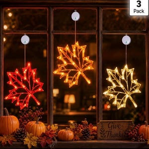 May include: A 3-pack of hanging maple leaf window decorations with integrated LED lights. The leaves are red, orange, and yellow, each suspended by a cord from a circular suction cup mount. The set is displayed in a window above pumpkins.