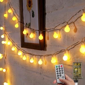 May include: A string of warm white LED globe lights with a thin wire, featuring a remote control with buttons for timer settings and brightness levels, and a standard electrical plug, creating a soft glow against a textured wall surface.