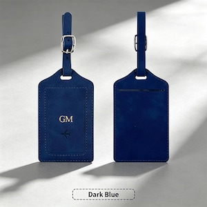 Puede incluir: Una etiqueta de equipaje azul oscuro con una correa ajustable a juego y una hebilla de metal plateado. El frente presenta las iniciales GM grabadas en oro sobre un pequeño icono de avión. El reverso muestra una ranura para tarjetas de identidad.