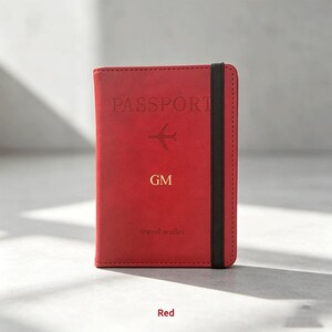 Puede incluir: Una cartera de viaje para pasaporte de color rojo con una banda elástica negra. La cubierta frontal tiene grabado el texto PASSPORT, un icono de avión, las letras GM en dorado y el texto travel wallet en una fuente pequeña y sutil.
