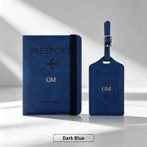 Puede incluir: Un portapasaportes azul oscuro con una banda elástica negra, que presenta el texto PASSPORT en relieve, un pequeño icono de avión y las iniciales doradas GM. Junto a él, una etiqueta de equipaje azul oscuro a juego con hebilla plateada.