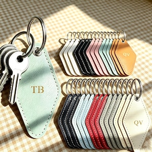 Puede incluir: Una colección de llaveros de piel sintética en forma de diamante en varios colores, incluyendo menta, blanco, negro, rosa, azul, rojo y tostado. Cada llavero tiene una anilla de metal y lleva grabadas iniciales como TB o QV en fuente serif.