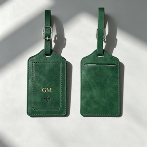 Puede incluir: Dos etiquetas de equipaje de color verde oscuro con correas ajustables y hebillas plateadas. Una etiqueta presenta las iniciales GM grabadas en oro y un pequeño icono de avión, mientras que la otra muestra una ranura horizontal para tarjetas.