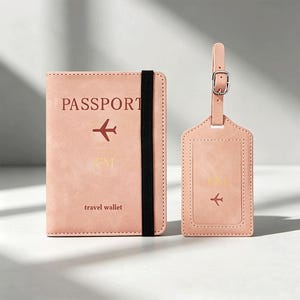 Puede incluir: Una funda para pasaporte de color rosa claro con una banda elástica negra, que incluye la palabra PASSPORT, un icono de avión, las letras GM y el texto travel wallet. Junto a ella hay una etiqueta de equipaje rosa a juego con hebilla.