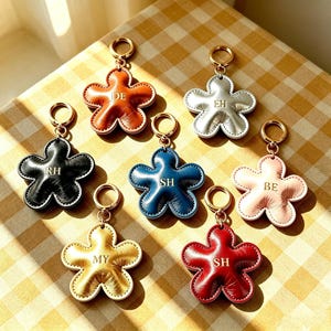 Puede incluir: Siete llaveros con forma de flor en varios colores, incluyendo marrón, plateado, negro, azul, rosa, dorado y rojo. Cada llavero cuenta con una anilla de metal dorado y está grabado con iniciales de dos letras: DE, EH, RH, SH, BE, MY, SH.