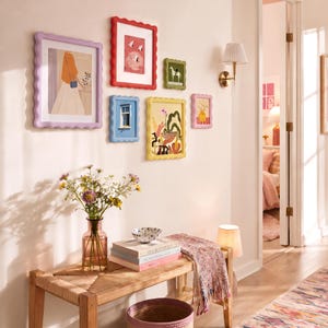 Puede incluir: Una colección de seis marcos de fotos coloridos con bordes ondulados montados en una pared, con varias láminas artísticas. Debajo, un banco de madera sostiene un jarrón de cristal con flores silvestres, un cuenco pequeño, libros y una manta.