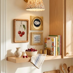 Op de afbeelding: Een houten plank bevat een keramische kom gevuld met rode kersen, een kleine bloemenvaas, een gevouwen gestreepte doek en een koperen boekensteun met kookboeken. Drie ingelijste kunstwerken tonen een kersenillustratie, een oester en drie vissen.