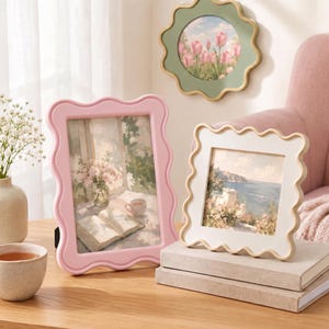 May include: A set of three wavy-edged picture frames including one large rectangular pink frame, one square cream frame, and one round sage green frame. Each frame contains a printed landscape or still life art piece with soft, muted color tones.