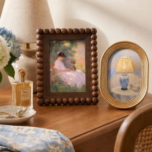 May include: A rectangular dark brown wooden picture frame with a beaded border containing an impressionist painting of a person and a dog, alongside an oval gold-toned ribbed picture frame featuring a painting of a lamp on a table.