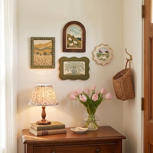 Pode incluir: Um candeeiro de mesa de madeira com um abajur plissado floral, dois livros empilhados, um vaso de vidro com tulipas cor-de-rosa e um pequeno prato. Na parede, quatro obras de arte emolduradas: uma moldura verde com contas, uma em arco, uma ondulada e uma festonada.