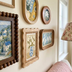 Pode incluir: Uma parede de galeria com várias peças de arte emolduradas, incluindo uma moldura de madeira retangular com borda recortada e acabamento em contas, envolvendo uma pintura de flores brancas numa jarra com padrão azul e branco sobre fundo bege.
