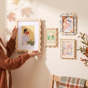 May include: Three rectangular wall frames with wavy edges. One large light wood frame holds a print of a person reading. A small light wood frame displays a green drink illustration labeled Spicy Marg. A floral patterned frame shows a food print.