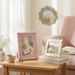 May include: A rectangular pink picture frame with a beaded border, a square white picture frame with a wavy edge, and a round green picture frame with a scalloped edge. The frames contain landscape and floral prints, displayed on a wooden table.