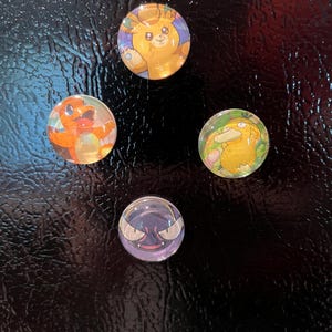 Könnte beinhalten: Vier runde, transparente Glasmagnete mit bunten Zeichentrickfiguren. Die Figuren sind ein Charmander, ein Psyduck, ein Gastly und ein bärenähnliches Wesen. Die Magnete sind vor einem schwarzen Hintergrund angeordnet.