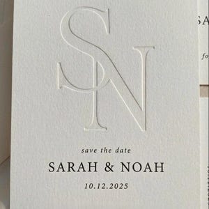 Op de afbeelding: Een witte kaart met de reliëfletters "S" en "N". De tekst "save the date" staat boven "SARAH & NOAH" en de datum "10.12.2025". Een minimalistisch ontwerp voor huwelijksaankondigingen. Bruiloftpapier.