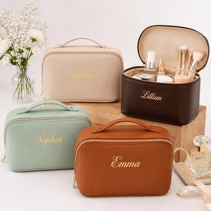 May include: Four rectangular cosmetic bags with top handles and gold zips. The bags are beige, dark brown, sage green, and tan, each featuring a name embroidered in gold script: Amanda, Lillian, Sophia, and Emma. The dark brown bag is open.