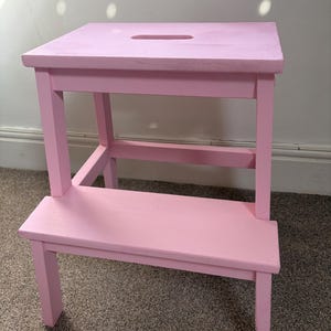 Puede incluir: Un taburete de madera rosa con una parte superior rectangular y un escalón inferior. La parte superior tiene un recorte de asa. El taburete está pintado de un color rosa claro sólido. Las patas son cuadradas y resistentes.