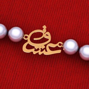 May include: A gold-colored pendant with Arabic script is attached to a pearl necklace. The pendant is centered on a red background. The pearls are large and white, and the script is in a stylized font.
