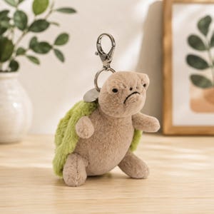 Puede incluir: Un llavero de tortuga de peluche con un cuerpo suave de color marrón claro y un caparazón texturizado de color verde brillante. La tortuga tiene una expresión facial gruñona con ojos pequeños negros y una boca hacia abajo. Incluye un mosquetón.