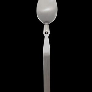 May include: A stainless steel spoon with a long, rectangular handle and an oval-shaped bowl. The spoon has a unique design with a small, decorative element connecting the bowl to the handle. The spoon is set against a black background.