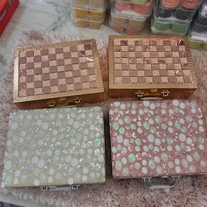 May include: Four decorative boxes with gold hardware. Two boxes have a checkerboard pattern in shades of gold and brown. The other two boxes are covered in iridescent sequins in shades of gold and pink. The boxes are arranged on a pink, textured surface.