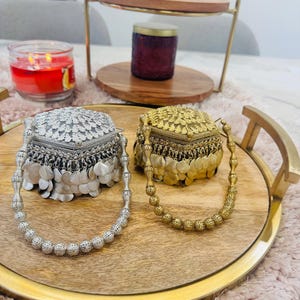 May include: Two hexagonal metal containers, one silver-toned and one gold-toned, each featuring an intricate filigree lid and a base adorned with dangling circular metal discs. Each container includes a matching beaded handle made of small spheres.